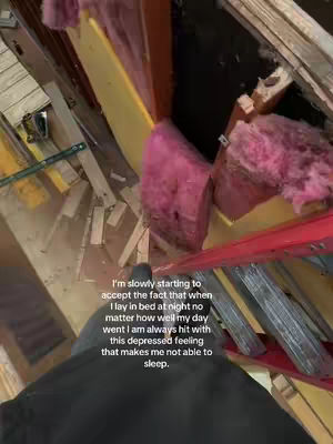 An interior view, likely of a house under construction or renovation, shot from a high angle, possibly from the attic or a higher level, looking down into the unfinished space. The scene includes exposed wooden beams, insulation materials, and various construction debris scattered around. The color palette incorporates the raw wood tones, the pinkish hue of the insulation, and other construction elements. The layout shows a structural framework, with areas appearing incomplete. A person's legs and part of their body can be seen, suggesting a first-person perspective. Text is overlaid on the image.