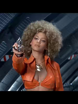 A still image of a woman holding a gun in a retro-futuristic setting. She has big, curly blonde hair and is wearing an orange outfit with a plunging neckline. The background has a science fiction aesthetic.