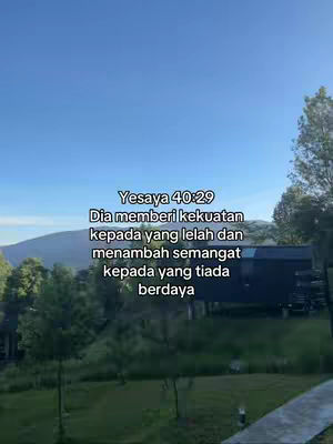 A scenic outdoor shot with a blue sky, green trees, and a building. The sky is a clear gradient from light blue at the bottom to a slightly deeper shade at the top. Trees and greenery frame the view. A dark building or structure is partially visible in the mid-ground, with a mountain range in the background . Text overlayed in the image shows a bible verse.