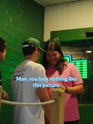 Two men are standing by a wooden barrier. A woman in a pink shirt stands behind with them. Text overlayed in white and blue reads "Man, you look nothing like this picture."