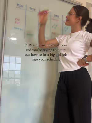 A young woman is standing in front of a whiteboard divided into columns labelled with days of the week, drawing on it. The whiteboard takes up most of the frame. The woman is wearing glasses, a white t-shirt and black pants, with her hair pulled back. Text overlayed reads "POV uni timetables are out and you're trying to figure out how to fit a big girl job into your schedule."