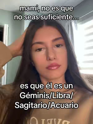 A woman is centered in the frame, her face is in focus. She has long, dark hair and is holding her hand near her head. Overlaid text at the top reads, 'mami, no es que no seas suficiente...' and text near the bottom reads, 'es que él es un Géminis/Libra/ Sagitario/Acuario'. The background is out of focus, with what seems to be a door and a curtain.