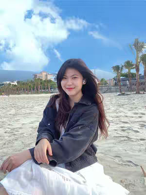 A young woman with long dark hair, wearing a black jacket over a white top and sitting on a sandy beach. The background includes a clear blue sky with scattered white clouds, palm trees, and buildings in the distance.