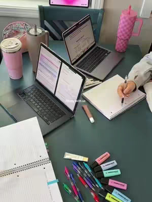 The image shows a student's study setup. Two laptops are open on a teal table, displaying study materials. A person's hand is writing in a notebook. Study supplies such as highlighters, pens, and lip balm are scattered around. Beverages and other study materials are on the table providing background.