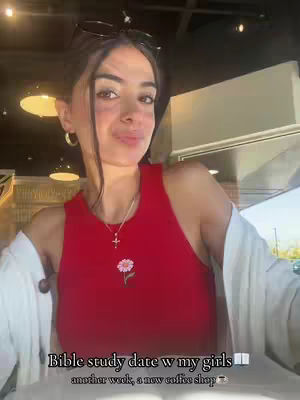 A selfie-style shot of a young woman indoors, likely in a coffee shop. She wears a red tank top and a white open shirt. Sunglasses are placed on her head. Text overlays on the bottom describe the content: 'Bible study date w my girls ... another week, a new coffee shop'. The lighting appears to be natural, contributing to a relaxed yet pleasant feel.