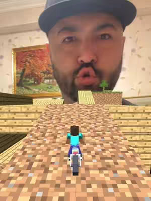 A man wearing a baseball cap is superimposed over a Minecraft scene displaying a dirt ramp, a Steve character on a motorcycle, and a small tree element. The man appears to be making a kissing face.