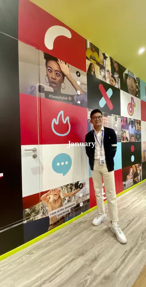 A full-body shot of a man standing against a wall decorated with various images and logos. He wears glasses, a black sweater, a white shirt, and light-colored pants. The word "January" is overlaid on the image.