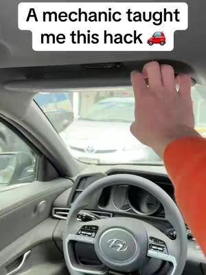 A person's arm and hand reaching up towards a car's sun visor, which is extended outwards. The interior of the car is visible, including the dashboard, steering wheel, and side mirror. In the background, another car is parked outside the window.