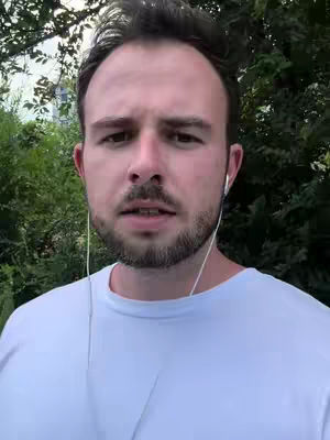 A close-up shot of a man looking directly at the camera with a serious expression. He is wearing a white t-shirt and earbuds are visible. The background is blurred greenery.