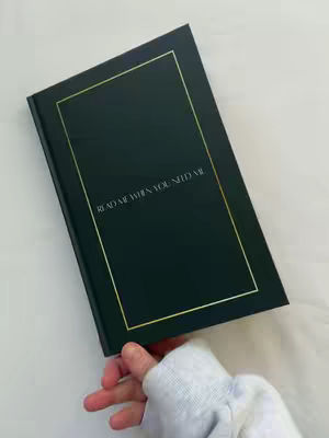 A hand holding a dark green book with gold trim and the text "READ ME WHEN YOU NEED ME" in the center. The background is white.