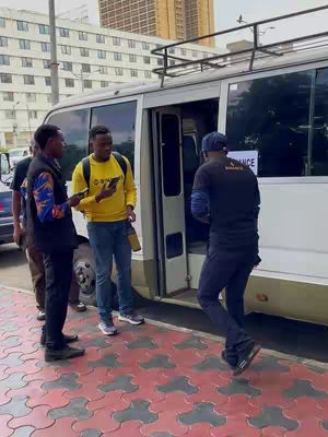 An outdoor shot with a white bus with its door open with several people standing around it, in what appears to be a city setting. The ground is covered in patterned paving stones, and in the background are buildings. The lighting appears to be daytime, with the sky not clearly visible. The general tone is active and candid.