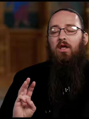 A close-up shot of a man with a full beard wearing glasses and a black shirt, raising his hand with two fingers extended. The background is slightly blurred, suggesting an indoor setting with warm lighting.