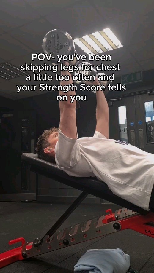 A man is lying on an inclined bench in a gym, performing a chest workout with dumbbells. The image is shot from a low angle, looking up at him. He wears a white t-shirt. Text overlayed on the image reads: "POV- you've been skipping legs for chest a little too often and your Strength Score tells on you". The background is dark with visible gym equipment.