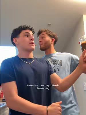 Two young men are holding hands, with one holding a glass of coffee near another's shoulder. The man closer to the camera is looking slightly to the side, and the man behind him is looking at him with a smile. They appear to be in a kitchen or apartment, as evidenced by the background.