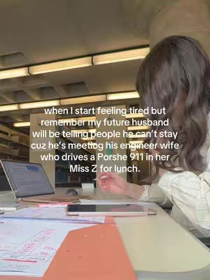 The image shows a person with brown hair, turned away from the camera, working on a laptop at a desk. The person is wearing a white shirt. There are papers and a tablet on the desk.  The background includes overhead lights and a glimpse of shelves.