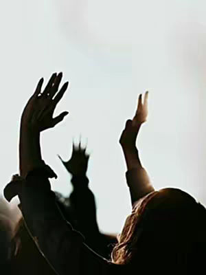 A silhouetted image of several people with their arms raised towards a bright light. The focus is on the hands, creating a sense of worship or celebration. The background is a bright, undetailed space possibly symbolizing sunlight or a light source. The color palette includes strong contrasts between the dark figures and the bright backdrop. The composition is vertical, emphasizing upward movement.