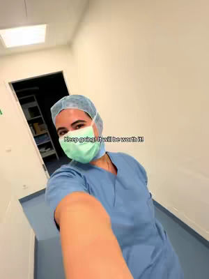 A selfie taken within a hospital hallway. The creator, a woman, is wearing a surgical cap, face mask, and blue scrubs. The background shows a clean, sterile environment with white walls, a visible door, and minimal equipment. The shot is framed from a first-person perspective.