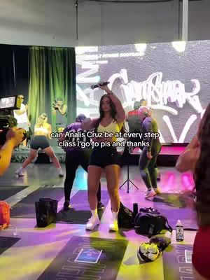 A woman in activewear stands center stage in a gym-like setting, holding a microphone aloft with her right hand, her left arm raised above her head. She is in a brightly lit room, with others in the background.