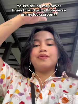 A young woman with dark hair and fair skin is the focus, wearing a white floral shirt and earphones. She is in a well-lit outdoor setting, with the sky and a metal structure visible in the background. Overlay text frames a question: "You're telling me I never knew I could put a Bible verse on my lockscreen??"