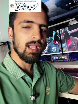 A man with a beard, wearing a green shirt, is speaking with his mouth open and tongue visible. Behind him is a computer screen displaying a video game character. The image is taken from a slightly low angle, emphasizing the creator's face and upper body.