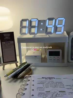 The image shows a tidy desk setup with a digital clock displaying 08:59, a small whiteboard with notes, several pens, and a notebook with handwritten notes. A small lamp provides soft lighting, and the overall aesthetic is minimalist and organized. The background includes a shelf holding a watch and a timer with text overlayed "Comment to stay on studytok".
