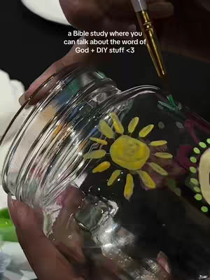 The image shows a close-up of a person painting a jar. The jar is clear glass and has been partially painted with a yellow sun design. The person is holding the jar with one hand and using a paintbrush with the other. The background is dark, and some text is overlaid on the image.