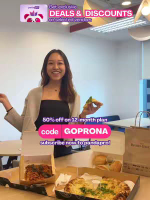A young woman smiles while holding a slice of pizza, with food items in boxes on a table in the foreground. The background features a modern office setting.