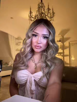 A woman with long blonde hair is centered. She is looking towards the camera with glossy lips and heavy makeup. She is wearing a white dress on the top, and there is a ornate chandelier above her head. The background shows a room with warm lighting, and another wall is visible on the left side of the camera.