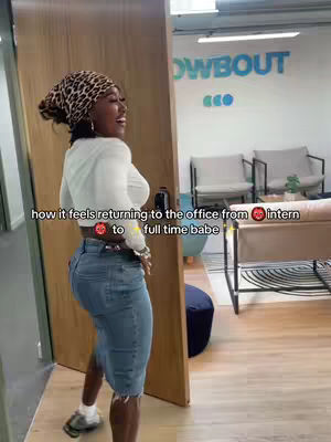 A woman in a casual outfit stands in an office setting, looking away from the camera and smiling. She wears a leopard-print headscarf, a white long-sleeved shirt, and denim shorts. The background includes doors, office furniture.