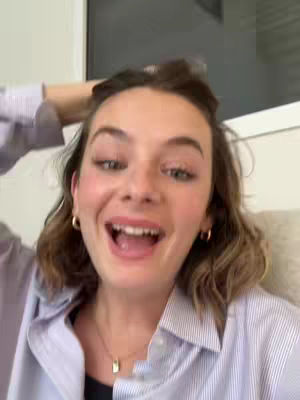 A woman with light skin and short, wavy brown hair is smiling at the camera, mouth open in a wide grin. She is wearing hoop earrings and a necklace with a rectangular pendant. Her shirt is a blue and white striped button down shirt. The background is a softly lit interior.