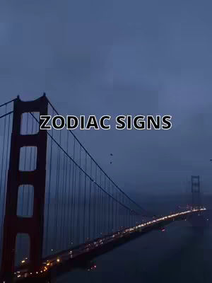 A wide shot of the Golden Gate Bridge at dusk, shrouded in a blue-grey mist. The bridge's red towers and cables are visible against the dim sky. The text overlayed reads 'ZODIAC SIGNS' in a white font.