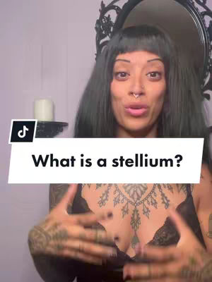 The cover image is a selfie-style shot featuring a woman in front of a wall. The woman has dark hair with a fringe, a nose ring, and tattoos on her arms and chest. She is in front of a distorted style design mirror, on an upper shelf there is a candle. Above the bottom section there is a text hook over a white block that says 'What is a stellium?'.