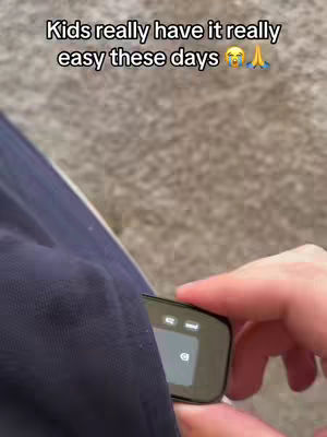 A gray carpeted background is visible. A navy blue garment extends from the top left corner, with an arm and hand holding a small, black, rectangular device that slightly protrudes from the fabric's edge. The device has a screen showing multiple icons, suggesting it's a piece of technology. The image features the text overlay, 'Kids really have it really easy these days' above the device and the hand.