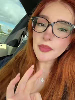 A close-up shot of a woman in a car. She has red hair and is wearing glasses. She is looking at the camera and smiling, with her fingers making a peace sign.