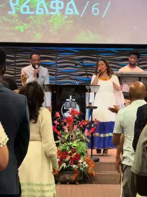 An image taken from a church service. The main focus is on a stage with a man and a woman singing into microphones and another man playing a keyboard in the background. A large floral arrangement sits in front of them.