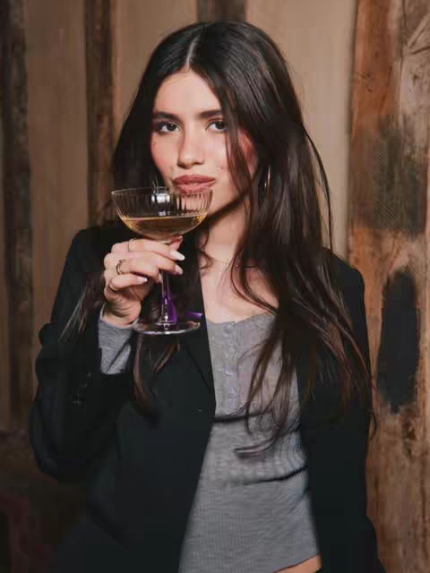 A woman with long dark hair is holding a champagne glass, looking at the camera with an upscale, after-dark vibe. She is wearing a dark blazer over a grey top. The background has a wooden panelled texture.