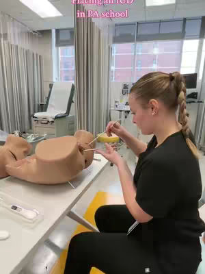 A woman is sitting at a table, a medical training mannequin in front of her. The room suggests a medical school setting, with medical equipment and a window visible. Overlay text suggests the topic is placing an IUD.