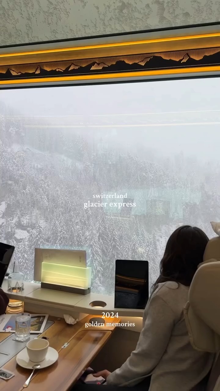 The image shows a person looking out of a train window, with a blurred, snowy mountain scene visible through the glass. Text overlays include 'switzerland glacier express' and '2024 golden memories'. The foreground features a table with a cup and saucer.