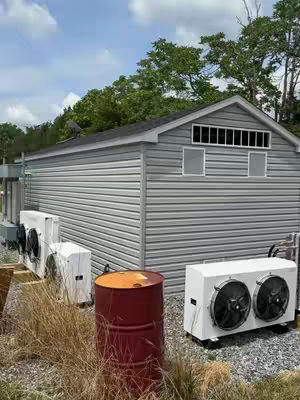 A low-angle shot of a gray shed with several white cooling units and a red barrel in front. The shed has a dark roof, and windows are visible. Tall grass and a blue sky with clouds in the background.