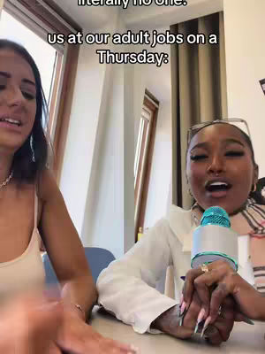 Two women are shown in a medium shot, both facing the viewer. The woman on the left is out of focus. The woman on the right is in sharp focus, holding a microphone, while looking to her right, and speaking. The background is a lightly colored well-lit space.