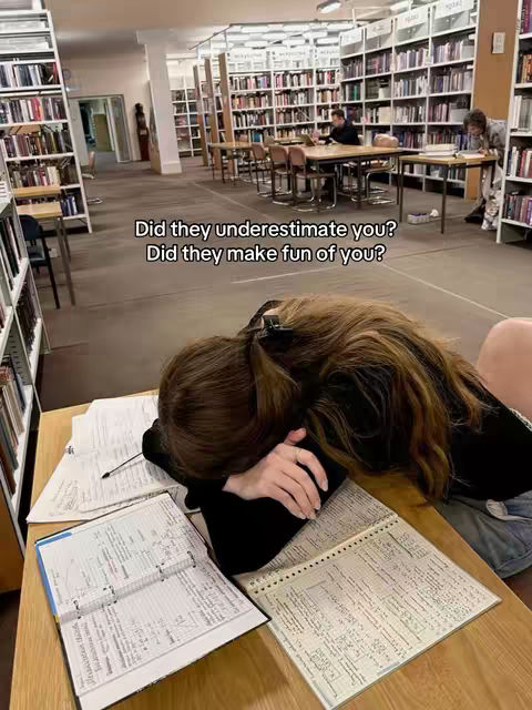 A young woman, seen from a medium angle, is resting her head and arms on a desk that holds books and papers. The scene takes place in a large library, with bookshelves lining the walls, and other people studying at nearby tables. The lighting is soft, creating an atmosphere of focused study and weariness.