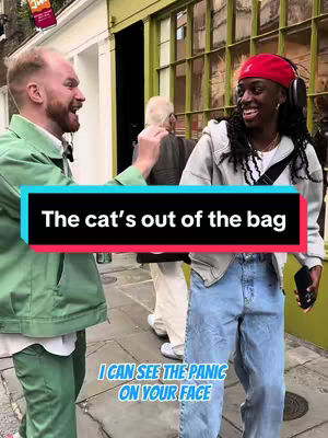 The cover image shows two people (one man, one woman) on a street, likely outside a shop judging by a partly visible store front. A text overlay with a blue and black background reading "The cat's out of the bag" is at the center, and another text overlay at the bottom saying "I CAN SEE THE PANIC ON YOUR FACE". The man on the left is wearing a green jacket and has his mouth open, while the woman on the right is wearing a red hat and is smiling. Behind them, another person is partially visible.