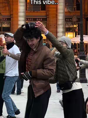 A group of people dancing in an outdoor setting, with a man in the center wearing a brown jacket and gesturing with his hands. A woman in a gray hat and jacket is dancing to the right. The scene suggests a lively and energetic atmosphere.