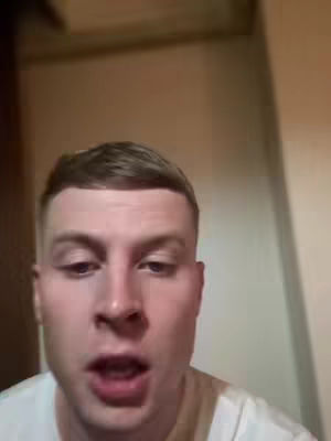 A close-up shot of a young man with short, light brown hair and a slightly open mouth, suggesting he is speaking. The background is out of focus, with glimpses of what appears to be a wall and a wooden surface, likely indoors. He is wearing a white t-shirt.
