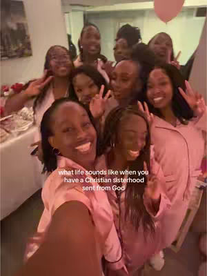 A group of young women are gathered closely together, smiling and posing for a selfie. They are indoors, and the lighting suggests a room with soft, warm illumination. The women are of various complexions, each wearing similar pink tops and some have matching robes. They are holding up peace signs and smiling joyfully.