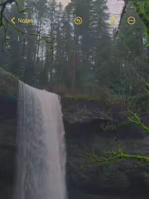 A scenic shot of a waterfall cascading down a rock face, surrounded by lush green trees. The image has a natural, misty quality, likely due to the presence of the waterfall. The composition is vertically oriented, emphasizing the height of the waterfall.