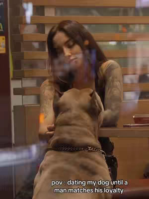 A woman with long brown hair is embracing a large dog, possibly a Cane Corso, in a restaurant setting. The woman has multiple tattoos on her arms, and the background includes wooden slat walls and visible glass reflections. The dog is wearing a chain collar.