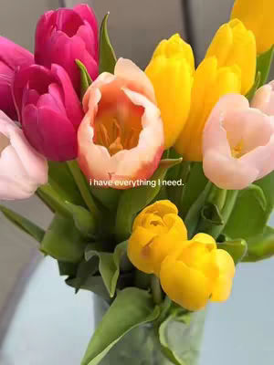 A vibrant bouquet of tulips in various colors, including pink, yellow, and orange, displayed in a clear vase against a soft, blurred background. The flowers fill the frame with their brightly colored petals.