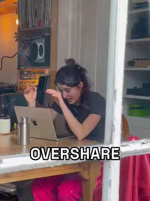 A woman sits at a wooden table in front of a laptop. She is leaning towards the laptop with an expression of surprise. Behind her is a window with curtains and shelves with books and other objects. The overlay text "OVERSHARE" is displayed in a white box at the bottom.