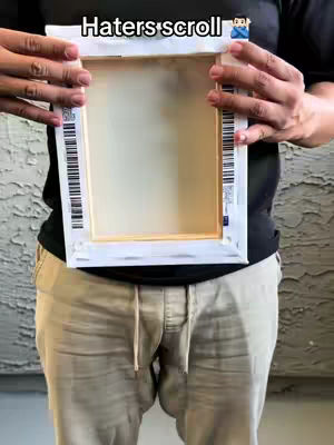 A person holds an empty canvas in front of their body, wearing casual clothing and standing in front of a textured wall. The canvas is rectangular, with a white frame and a blank, light-colored center. The person's hands are visible, holding the canvas at the top and bottom sides. The background features a patterned wall, and the overall composition is casual and straightforward, with the focus on the empty canvas.